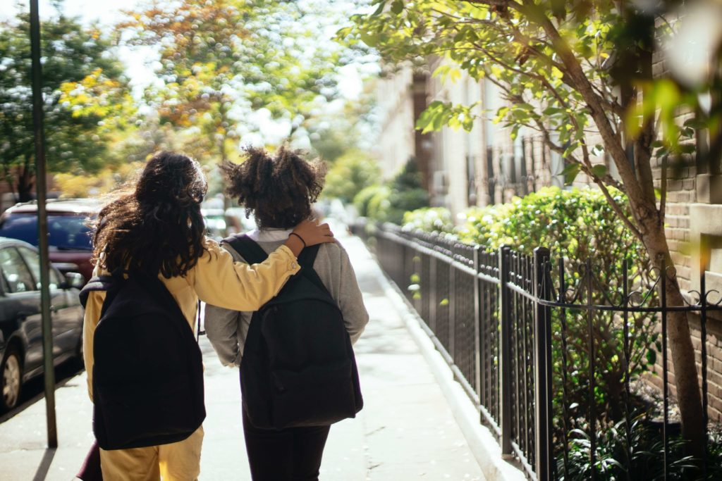 Image of two girls with backpacks walking down the sidewalk with their backs to the camera. Montessori preschool